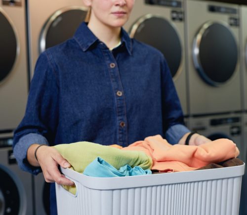 Cropped shot of girl holding basket with load of laundry preparing colored clothes for washing at self service laundromat, copy space